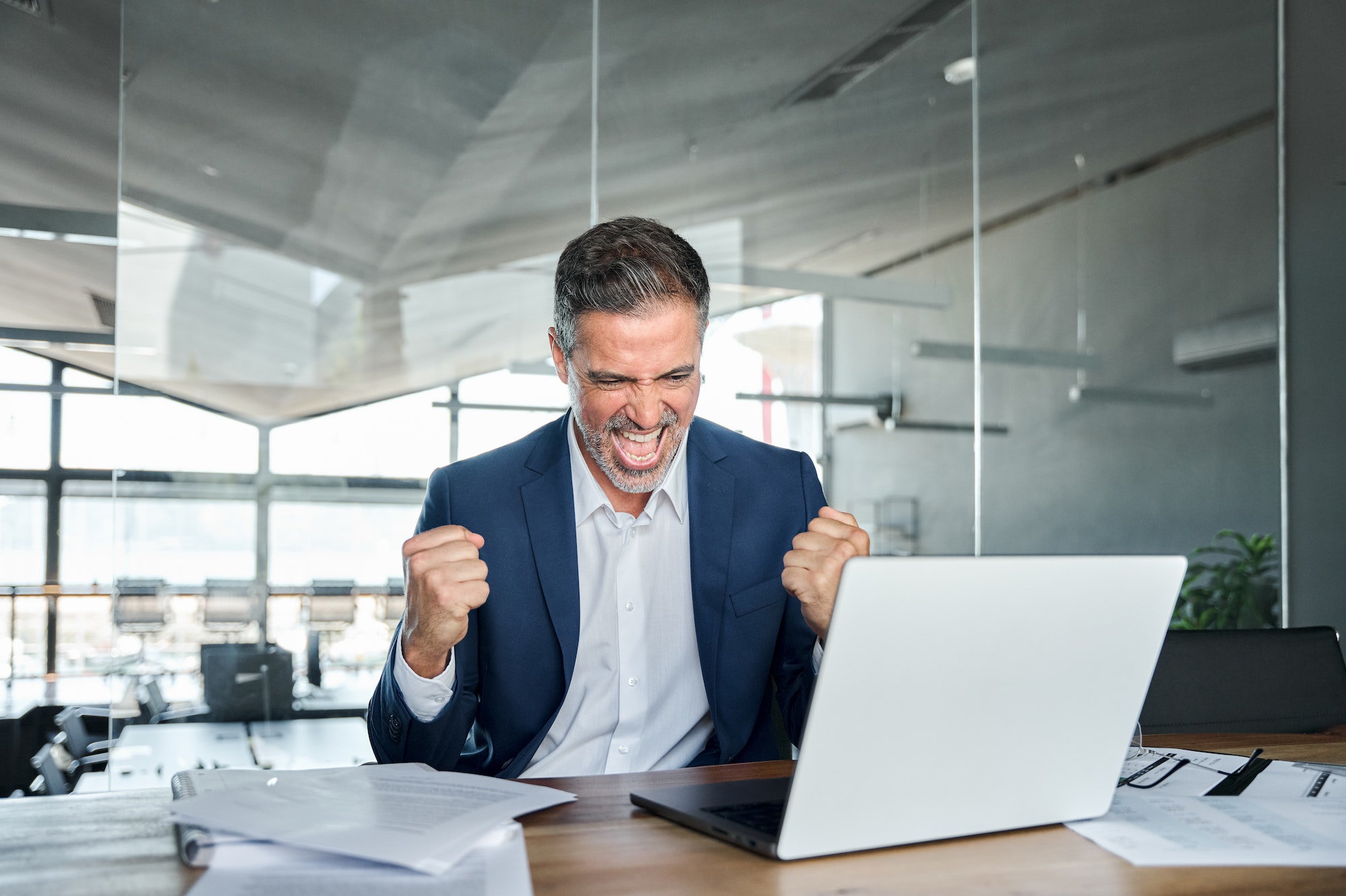 Happy mid aged business man celebrating success in office looking at laptop.